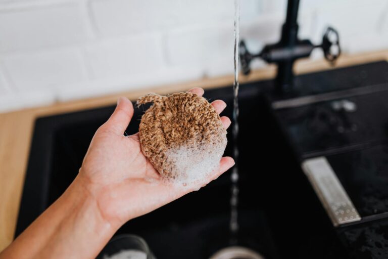 Close-up of a hand holding a natural sponge under running water at a sink.